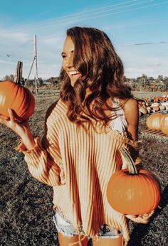 a woman holding two pumpkins in one hand and smiling at the camera while standing in an open field