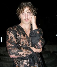 a young man with curly hair wearing a black lace shirt and jeans posing for the camera