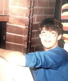 a young man wearing glasses sitting in front of a fire place with his arm out