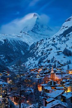 a snowy mountain with houses in the foreground and lights lit up on the buildings below