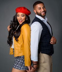 a man standing next to a woman in front of a gray background wearing a red hat