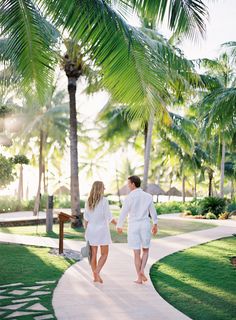 a man and woman walking down a path in the grass with palm trees behind them