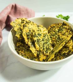 there is a white bowl filled with spinach chips on the table next to a napkin