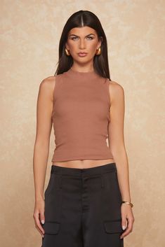 A woman with long dark hair stands against a textured beige background. She is wearing the Talia Ribbed Tank - Java by Noli Yoga and black high-waisted pants, creating a flattering slim silhouette. The ultra-soft rib material of her fitted brown top complements her accessories: large hoop earrings and a gold bracelet, as she looks directly at the camera. Brown Ribbed Sleeveless Top, Brown Seamless Sleeveless Top, Solid Seamless Tank Top With 4-way Stretch, Non-stretch Ribbed Sleeveless Tops, Black High-stretch Yoga Tank Top, Sleeveless Mock Neck, Mock Neck Top, Ribbed Tank, Staple Pieces