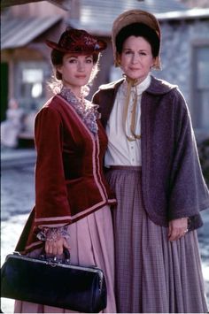 two women standing next to each other in long dresses and hats, one holding a purse