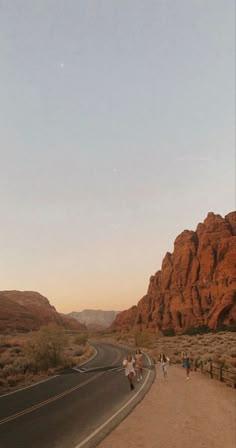 people are walking down the road in front of some red rocks and mountains at sunset