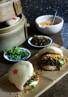 two buns with meat and vegetables on a cutting board next to bowls of food