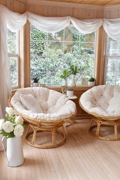 two wicker chairs in front of a window with white curtains and flowers on the table