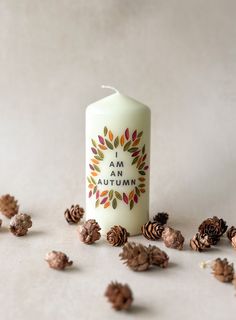 a white candle with an i am autumn sticker surrounded by pine cones