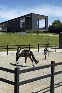 a woman walking behind a horse in an enclosed area with a black fence and building