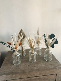 three clear vases with dried plants in them on a wooden table next to a white wall