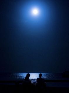 two people sitting on a bench in front of the ocean under a full moon at night