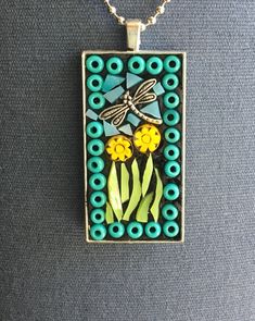 a necklace with a dragonfly sitting on top of yellow flowers and green leaves in front of a gray background