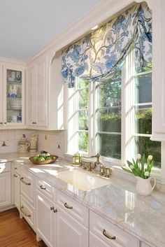 a kitchen with white cabinets and marble counter tops in front of a window that has blue floral curtains