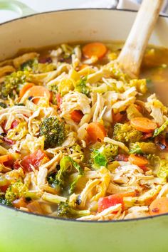 a green pot filled with chicken, broccoli and carrots next to a wooden spoon