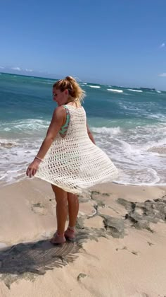 a woman standing on top of a sandy beach next to the ocean in a white crocheted dress