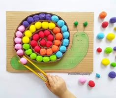 a child's hand holding a pair of scissors next to a paper plate with a snail made out of pom - poms