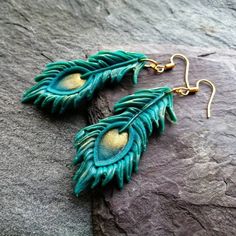 pair of peacock feather earrings with gold accents on top of a stone slab in front of a rock