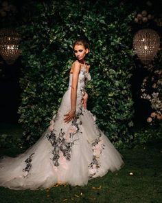 a woman standing in front of a lush green wall wearing a wedding dress with flowers on it