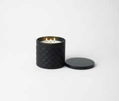 a candle that is sitting next to a black container on a white surface with two candles in it