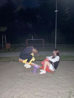 two people sitting in the sand at night with skateboards and one person holding a cell phone