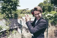 a man in a black jacket holding a red rose up to his face while standing next to a fence