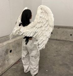 a woman with white wings on her back standing in front of a concrete wall and looking down at the ground