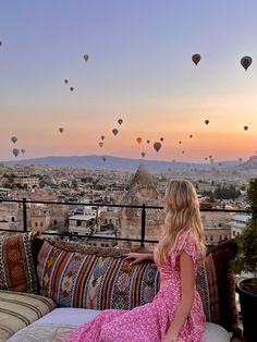 a woman in a pink dress sitting on a couch looking at hot air balloons flying over the city