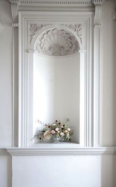 a white fireplace mantel with flowers on top and an arch above it in the corner