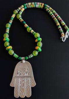 Sterling Silver (acid tested) 'Hamsa' hand-decorated with fine geometric designs. The Hamsa or Hand of Fatima is a palm-shaped amulet of an open right hand recognized (since ancient Egypt) as a sign of protection against evil. Strung on necklace with pretty green reconstituted stone, recycled green glass, and multi-color 'Bakelite' style disc beads. Finished with a contemporary silver plated closure making a lovely vibrant good luck charm. *PLEASE NOTE THAT YOUR PHONE NUMBER IS REQUIRED SEPARATELY TO INITIATE SHIPPING* Traditional Green Engraved Necklace, Green Amulet Jewelry For Festivals, Green Amulet Jewelry For Festival, Bohemian Green Engraved Necklace, Protection Against Evil, Snowflake Necklace, Hamsa Necklace, Friendship Jewelry, Luck Charm