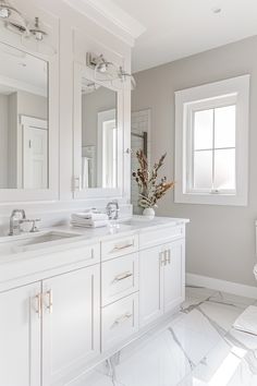 a white bathroom with two sinks, mirrors and a toilet in the corner next to a window