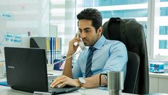 a man sitting at a desk with a laptop and cell phone in his hand while talking on the phone