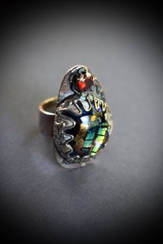 This beautiful silver ring is form by freeform organic shapes and lines with beautiful green and gold dichroic cabochon in the center and red cubic cz on top. It have been oxidized to bring out the texture with a slightly colored patina. I polished it to a shine, but left the patina in the recesses to bring out the texture. It's a perfect ring for everyday wear and has a rustic artisan quality. Dimensions: ring size 8. 100% eco friendly, recycled silver. Ready to ship! Your purchase will arrive Green Metal Cabochon Jewelry, Green Opal Cabochon Ring, Fordite Ring, Unique Cabochon Emerald Ring, Collectible, Bohemian Green Beads, Gems, And Cabochons, Recycled Silver, Dichroic Glass, Perfect Ring, Organic Shapes