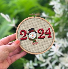 a person holding up a cross - stitch christmas ornament in front of some flowers