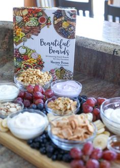 a wooden tray topped with lots of fruit and nuts next to a book on top of a table