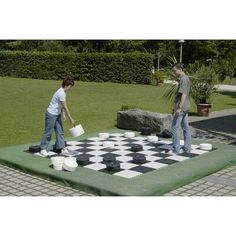 two people playing checkers on a giant chess board in a park area with grass and bushes