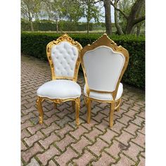 two white and gold chairs sitting next to each other on a brick walkway with hedges in the background