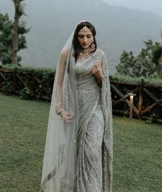 a woman in a white sari and veil standing on grass with mountains in the background