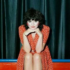 a woman sitting on top of a red bench in front of a black curtain with her legs crossed