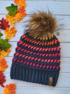 a knitted hat with a pom - pom sits next to fall leaves