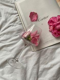 pink flowers are placed in a wine glass next to an open book on a bed