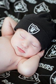 a baby wearing a oakland football beanie laying on a blanket with his eyes closed