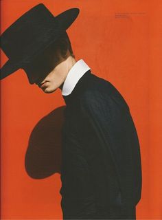 a man with a black hat and shirt on standing in front of a red wall