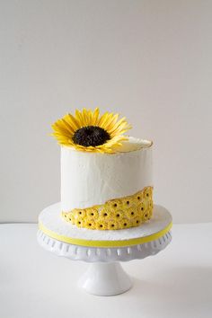 a white cake with yellow decorations and a single sunflower on the top, sitting on a pedestal