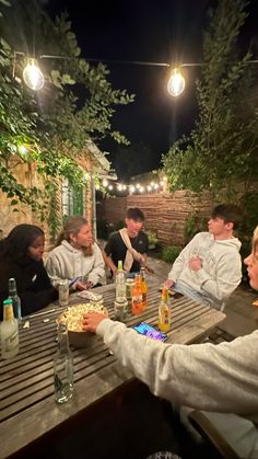 a group of people sitting around a wooden table eating food and drinking beer at night