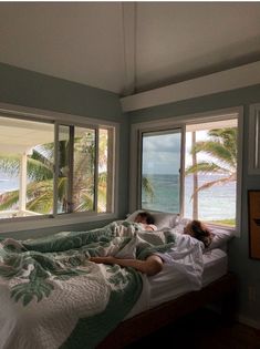 two people laying in bed looking out the window at the ocean and palm trees outside