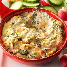 a red bowl filled with cheese and vegetables next to sliced cucumbers on a white surface