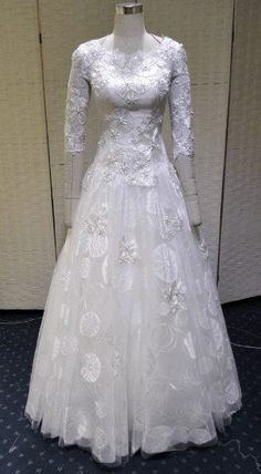 a white wedding dress with long sleeves and flowers on the skirt is displayed in front of a wall