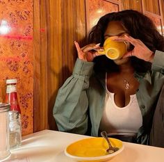 a woman sitting at a table drinking from a yellow cup with her hands up to her face