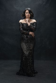 a woman in an off the shoulder black dress posing for a studio photo with her hands on her chin